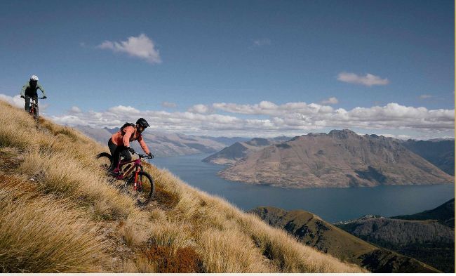 Two mountain bikers descending ridge with Lake Wakatipu and mountains visible