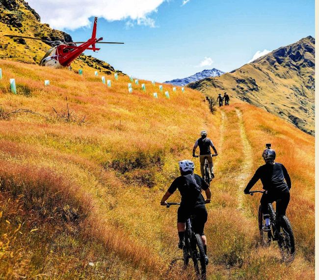 Three mountain bikers riding uphill trail with red helicopter above