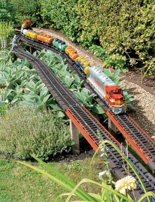 A Santa Fe SD45 diesel locomotive numbered 5610 in warbonnet livery leads a long train of colourful tank cars across a timber trestle bridge through a lush garden landscape.