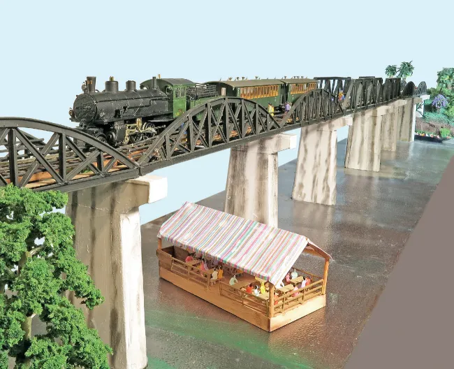 A detailed model railway layout depicting a stone viaduct with a steam locomotive and rolling stock passing through a scenic rural setting.