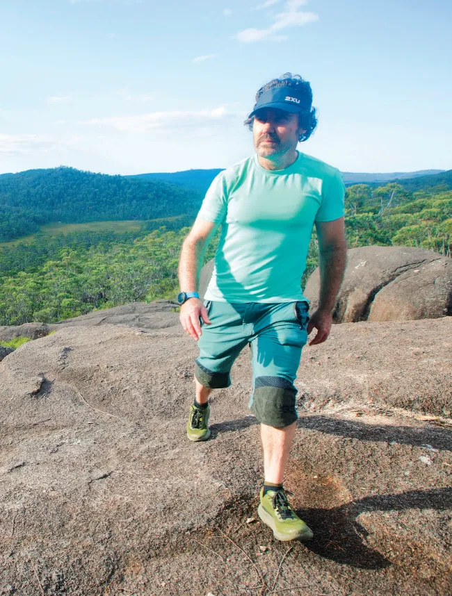 Hiker in a teal shirt and shorts walking across open granite slabs with forested valleys stretching to the horizon behind him.