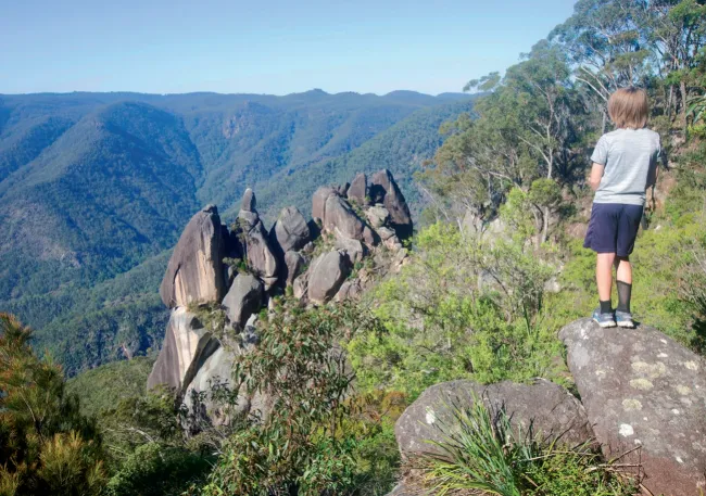 A child stands on a granite outcrop gazing out over a sweeping valley with dramatic rocky pinnacles and forested ridges below.